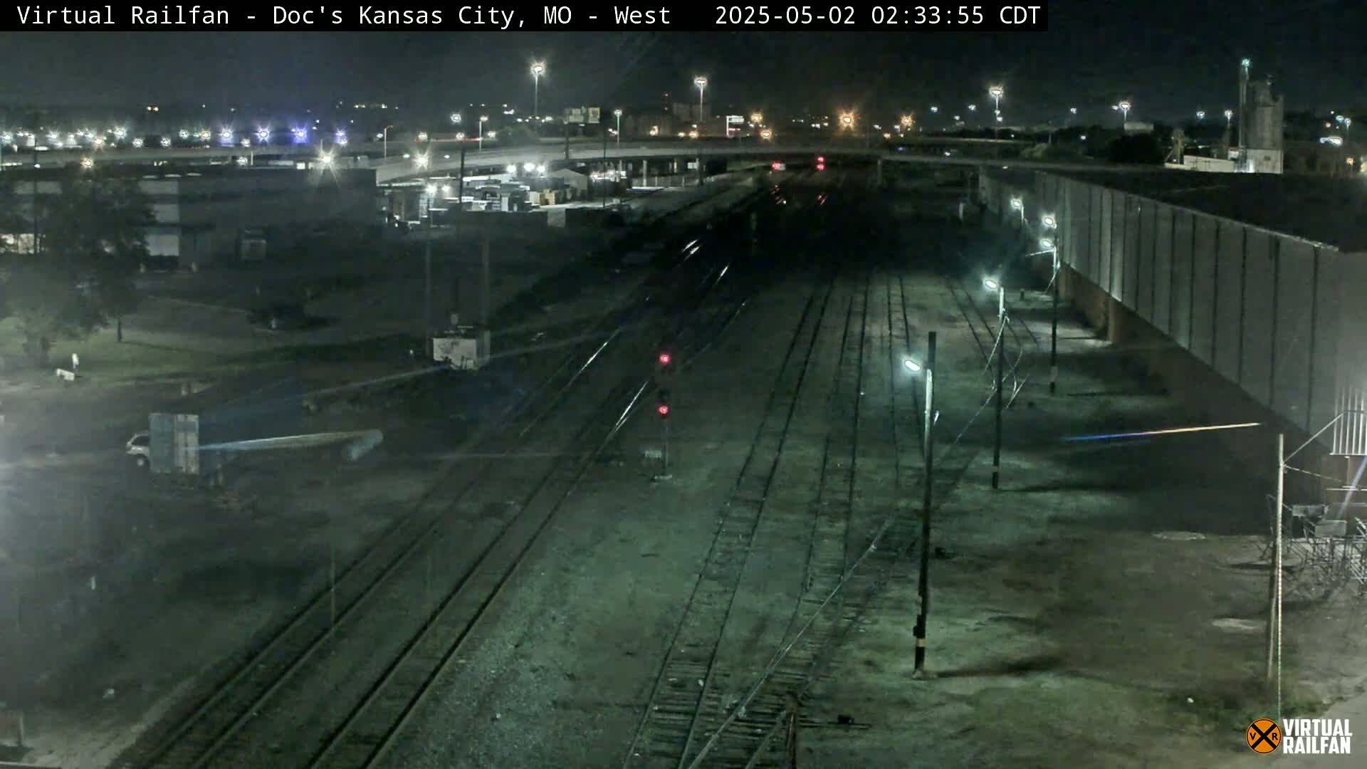 A nighttime view of railroad tracks stretches into the distance, passing by industrial buildings and a highway overpass under a clear sky.