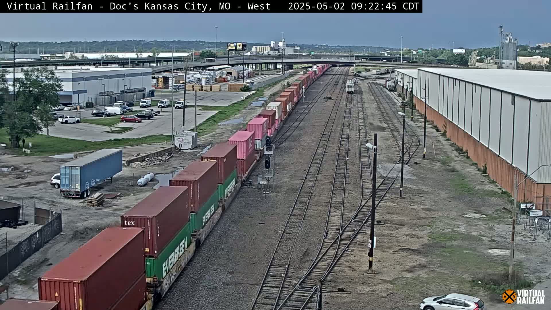 A long freight train carrying various colored shipping containers sits on railroad tracks next to industrial buildings under an overcast sky.
