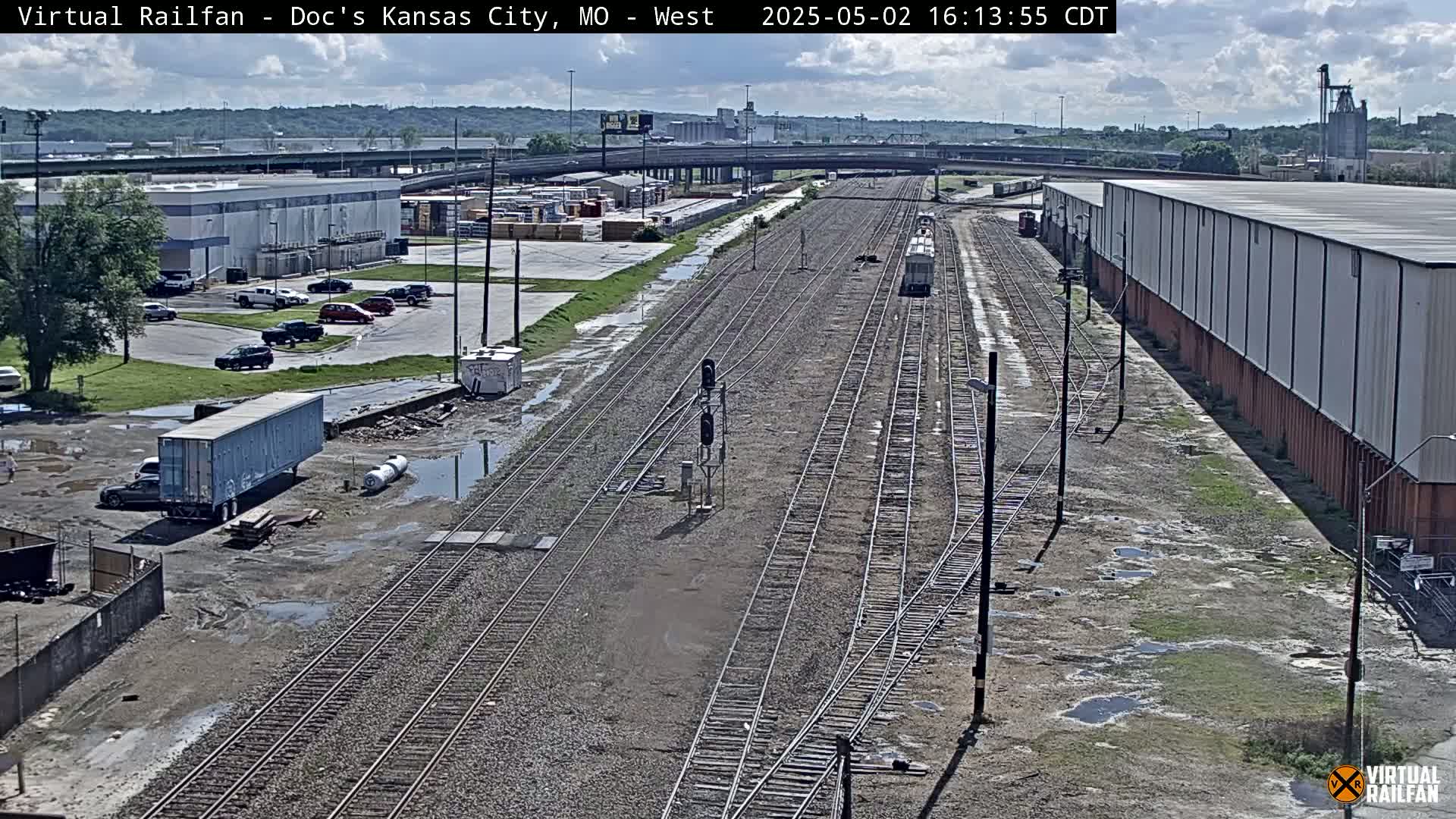 A mostly sunny day shows a view of railroad tracks splitting into multiple directions, surrounded by industrial buildings, parking lots with cars, and several puddles in a mostly empty lot.