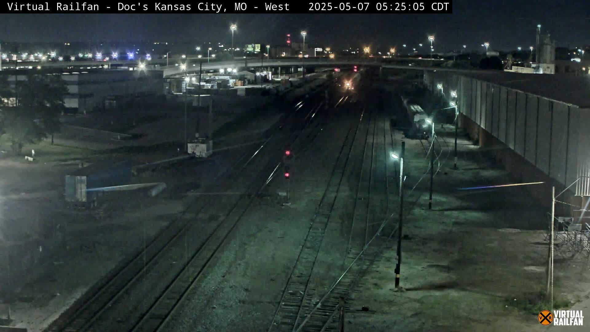 A nighttime view of a train yard with multiple sets of train tracks, some railway cars, and industrial buildings under a clear, dark sky.