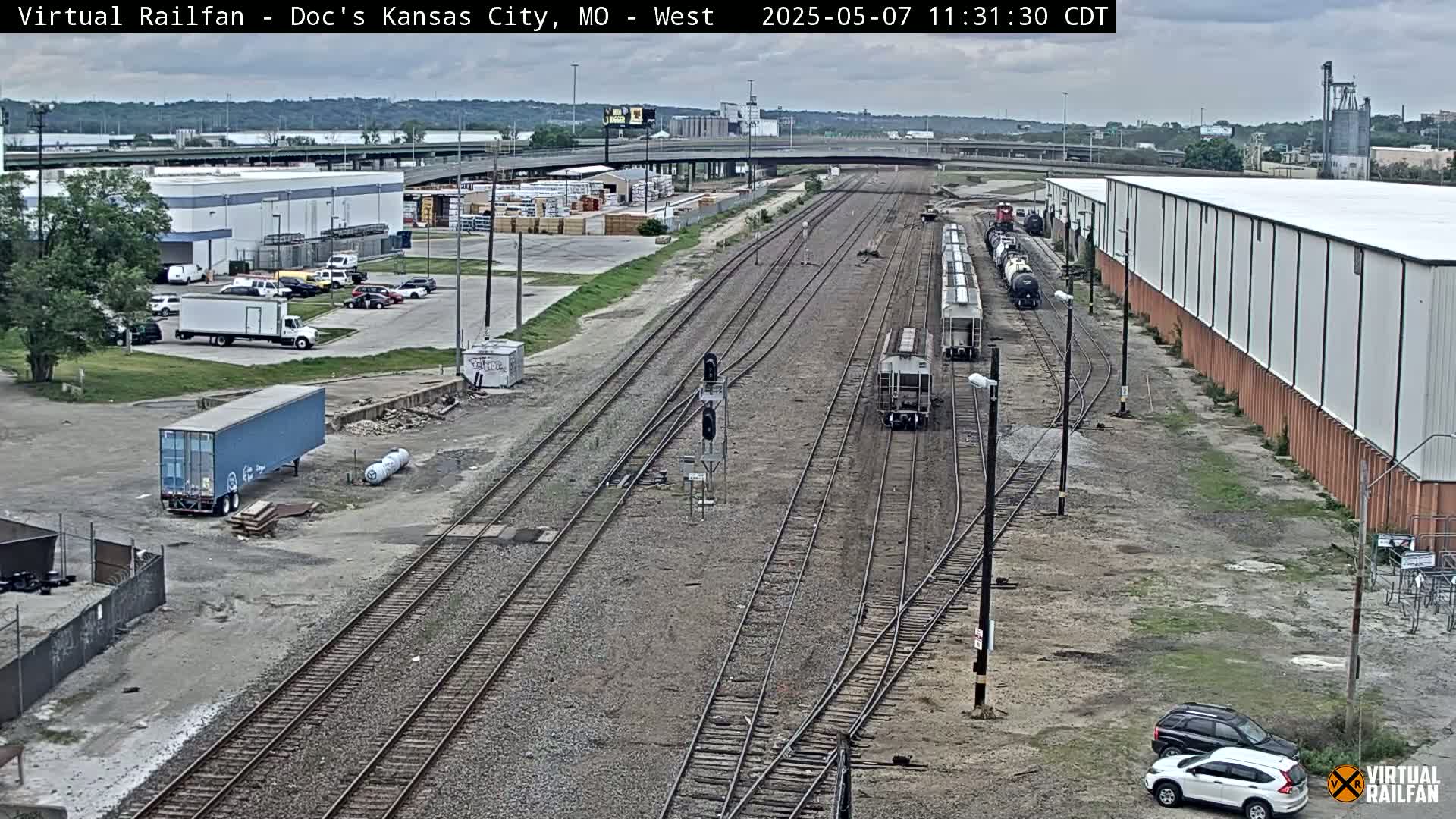 A high-angle, long shot of a railway yard with several train cars, surrounded by industrial buildings and parked cars under an overcast sky.