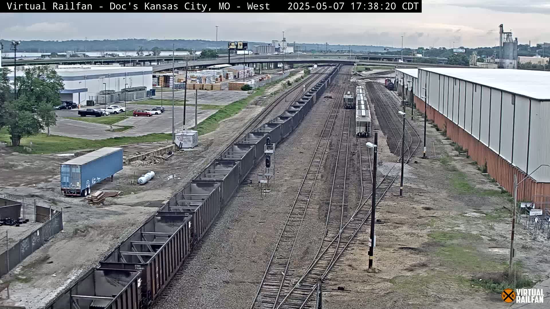 A long freight train carrying dark cargo sits on railroad tracks near industrial buildings and parking lots under an overcast sky.