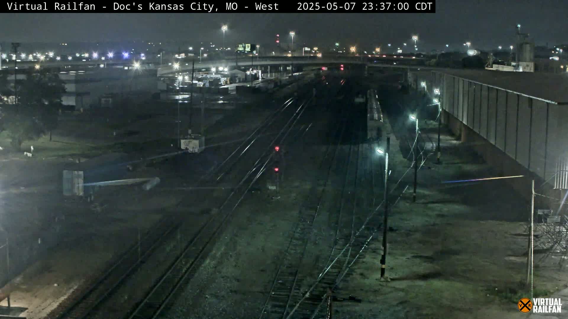 A nighttime, overhead view of a train yard with multiple sets of train tracks, some freight cars, and surrounding industrial buildings under artificial light.