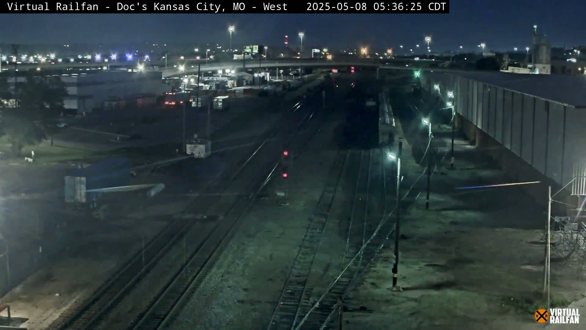 A nighttime view of a railroad yard with several train tracks, some industrial buildings, and vehicles, under a clear, dark sky.