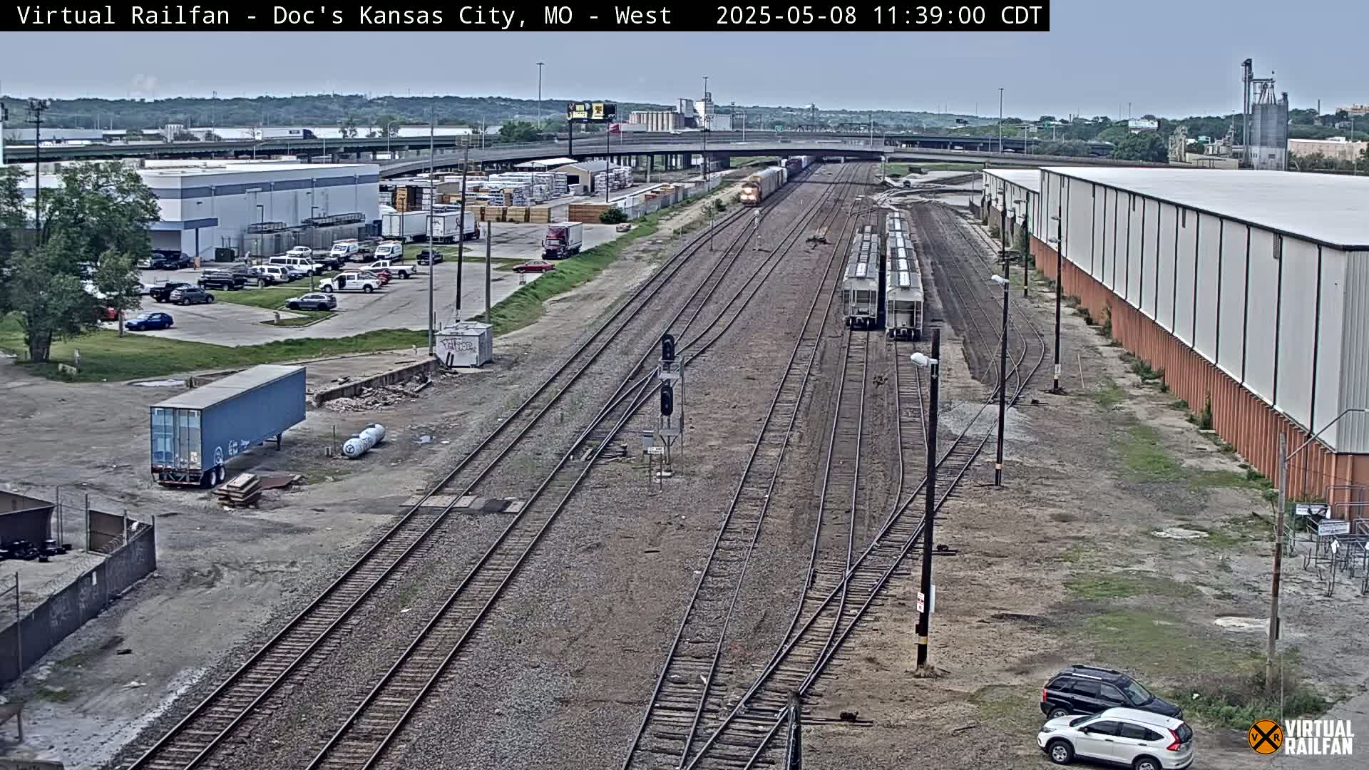 A high-angle, long shot shows several train cars on railway tracks near a warehouse and parking lot under a partly cloudy sky.