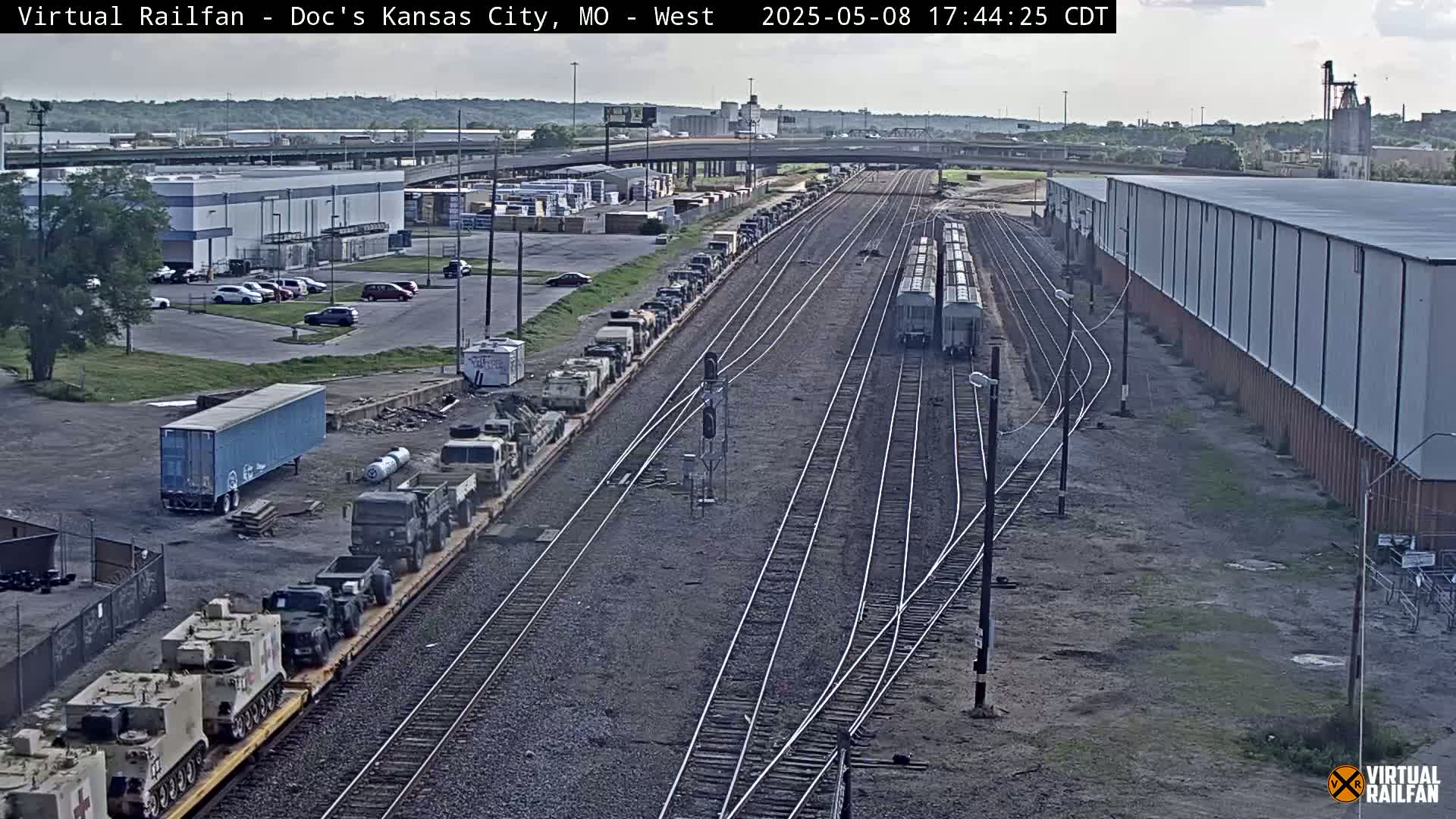 A long freight train carrying military vehicles sits on a railway next to industrial buildings under a partly cloudy sky.