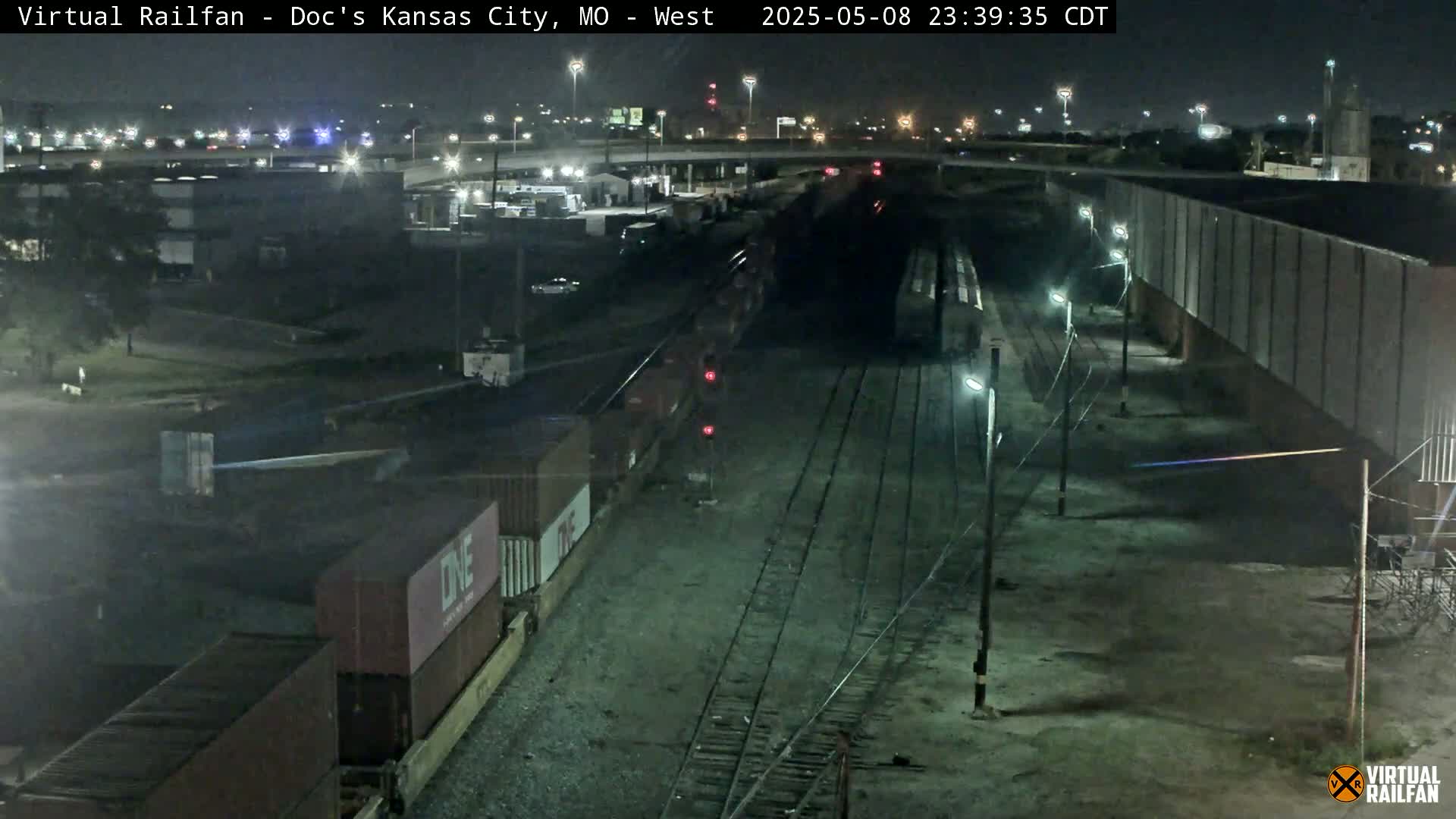 A nighttime view of a freight train alongside railroad tracks near industrial buildings under a clear sky.