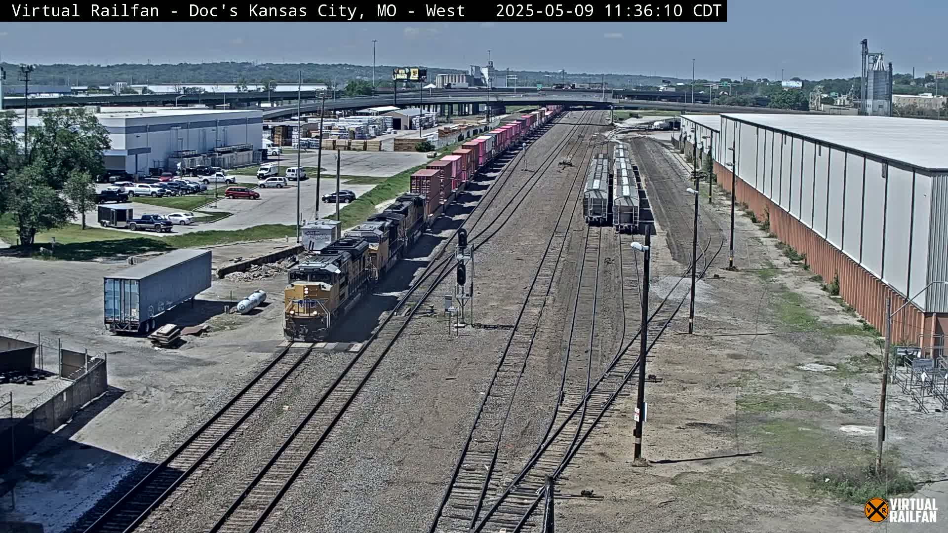 A long freight train, composed of multiple locomotives and numerous cargo containers, is situated on railroad tracks beside a parking lot and industrial buildings under a mostly sunny sky.