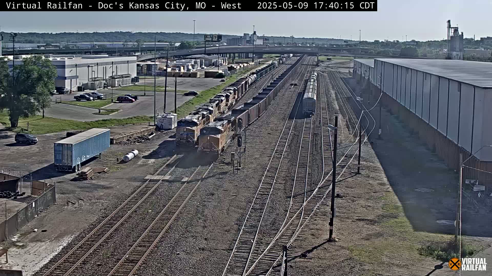 A long freight train passes through a rail yard on a sunny day, with other trains and industrial buildings visible in the background.