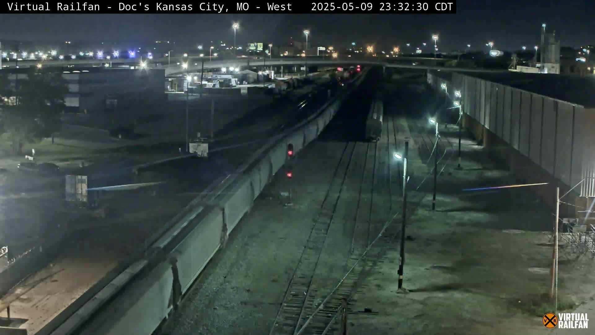 A long freight train moves along a set of railway tracks at night, passing under a highway overpass and alongside industrial buildings.