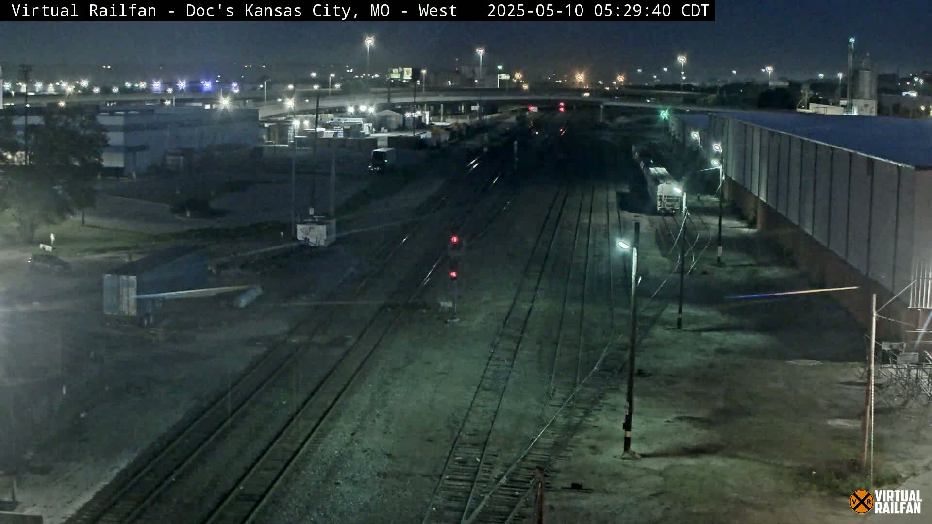 A nighttime view of a railway yard with several train cars and tracks, industrial buildings, and a highway overpass in the distance.