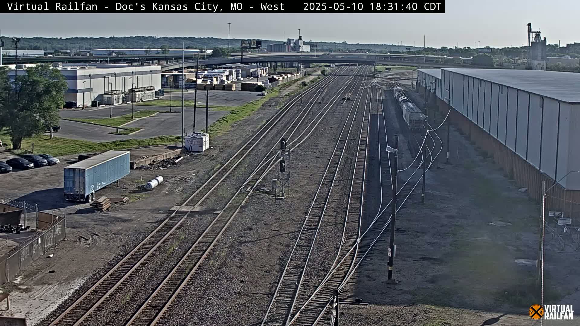 A long freight train sits alongside several sets of railroad tracks near industrial buildings and parking lots on a sunny day.