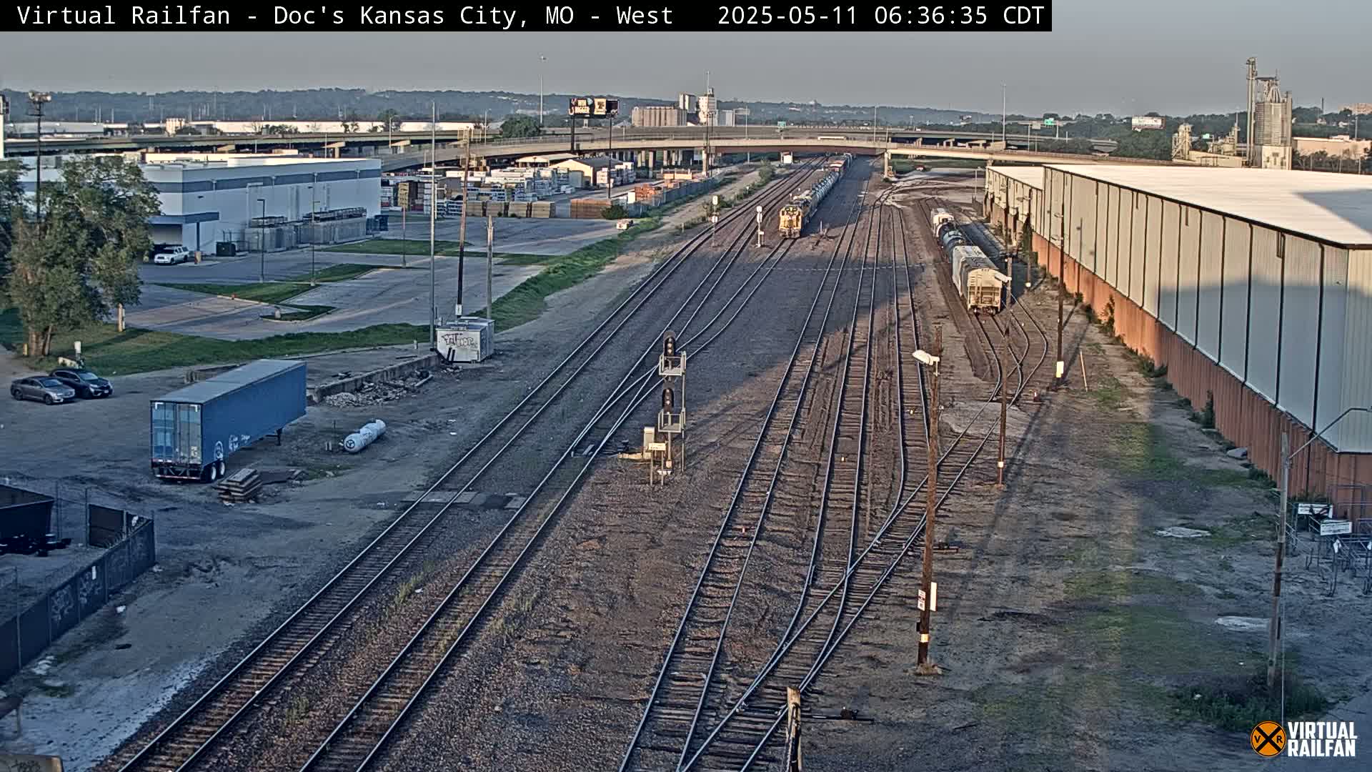 A high-angle, long shot of a train yard with several train cars and a few trucks under a partly sunny sky.