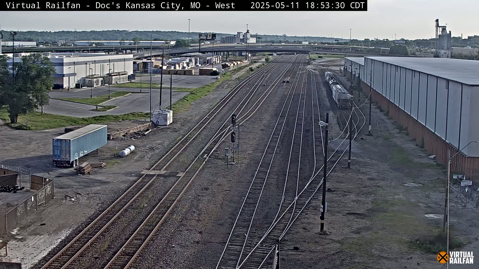 A mostly clear day reveals a sprawling rail yard with multiple train tracks, industrial buildings, and a lone freight train, a semi-trailer truck, and other vehicles.