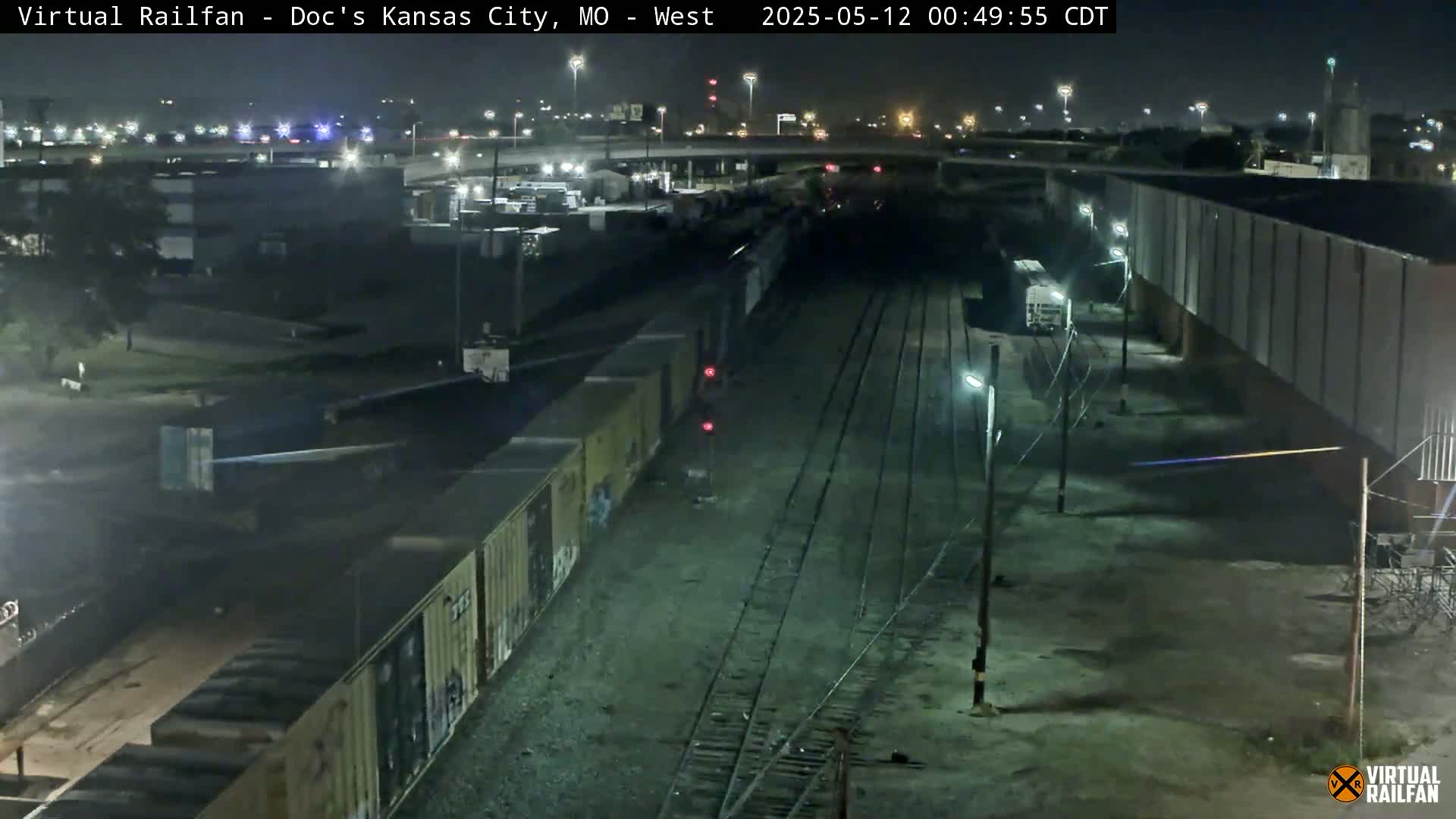A nighttime view of a train yard with a long freight train and a few other vehicles parked alongside railroad tracks under artificial lights.