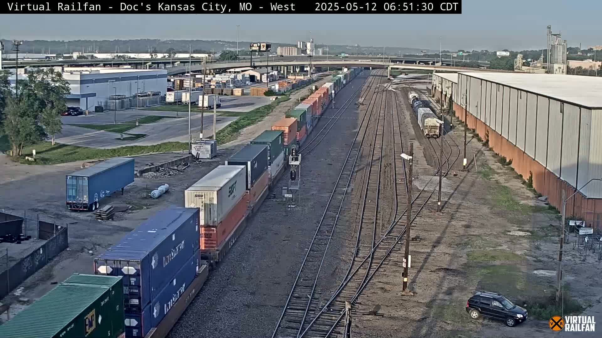 A long freight train carrying multiple shipping containers is visible on railroad tracks near industrial buildings under a clear sky.