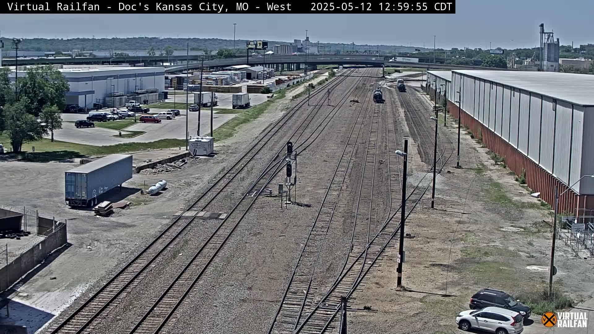 A sunny view of railroad tracks intersecting with a roadway, surrounded by industrial buildings and parked cars.