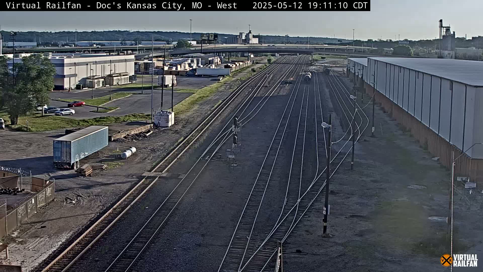 A high-angle, long shot of a railway yard on a partly cloudy day, showing several train tracks, industrial buildings, and a few vehicles.