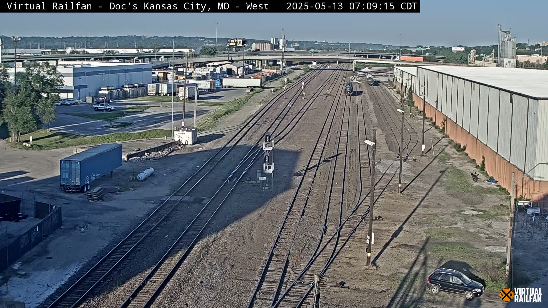 A high-angle view of a train yard with multiple train tracks, industrial buildings, and a few vehicles, on a sunny day.