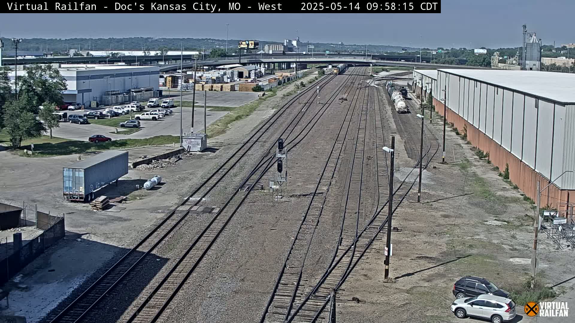 A high-angle view of railroad tracks on a sunny day, with industrial buildings, parked cars, and a freight train visible.