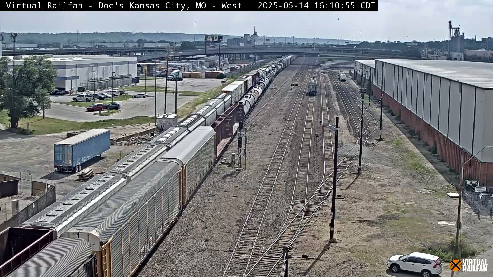 A long freight train sits on railroad tracks next to industrial buildings on a sunny day.
