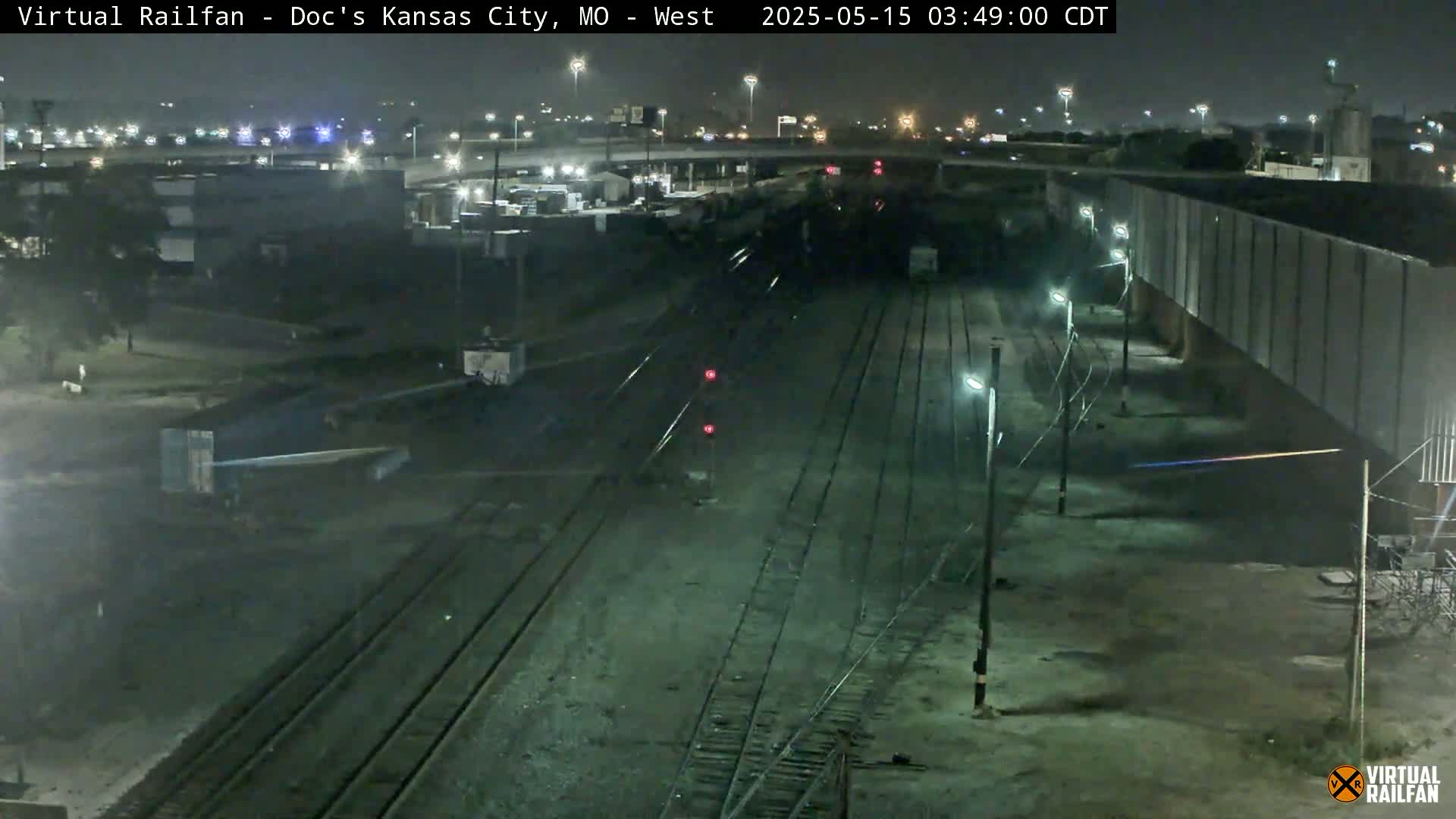 A nighttime view of a train yard with multiple sets of railroad tracks, surrounded by industrial buildings and illuminated by streetlights.