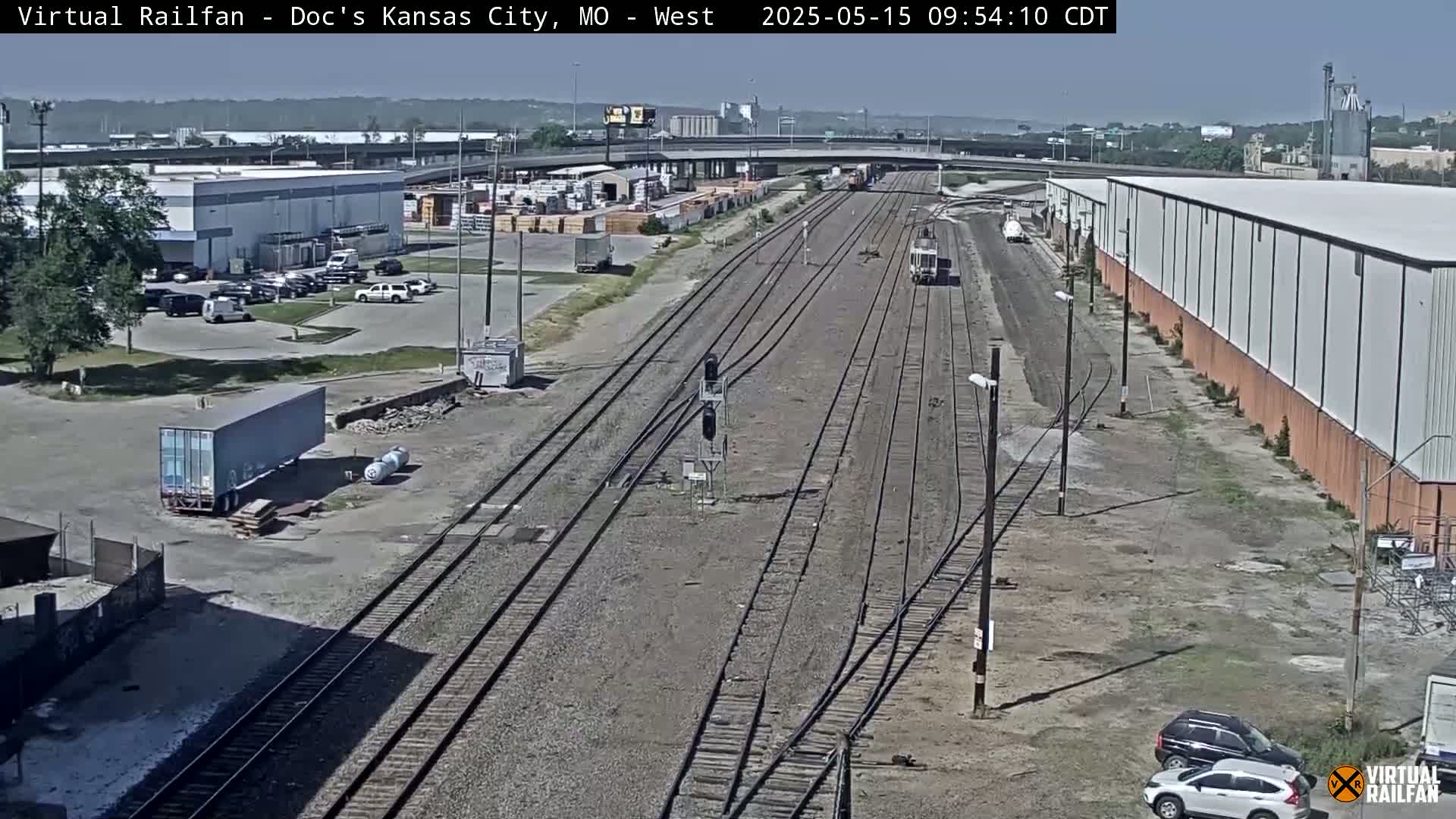 A high-angle, long shot shows several railroad tracks stretching into the distance on a sunny day, surrounded by industrial buildings, parking lots, and some vehicles.