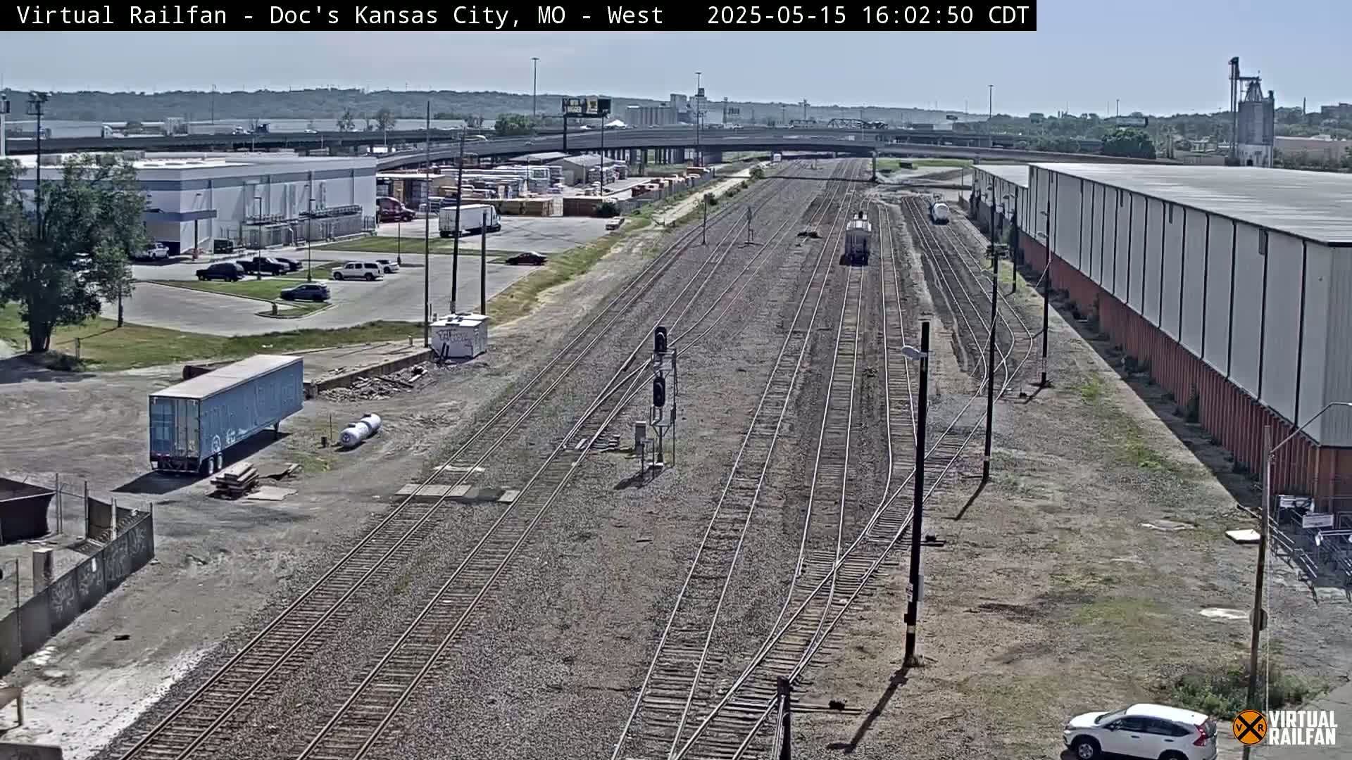 A long stretch of railroad tracks stretches into the distance under a clear sky, with industrial buildings and vehicles visible on either side.