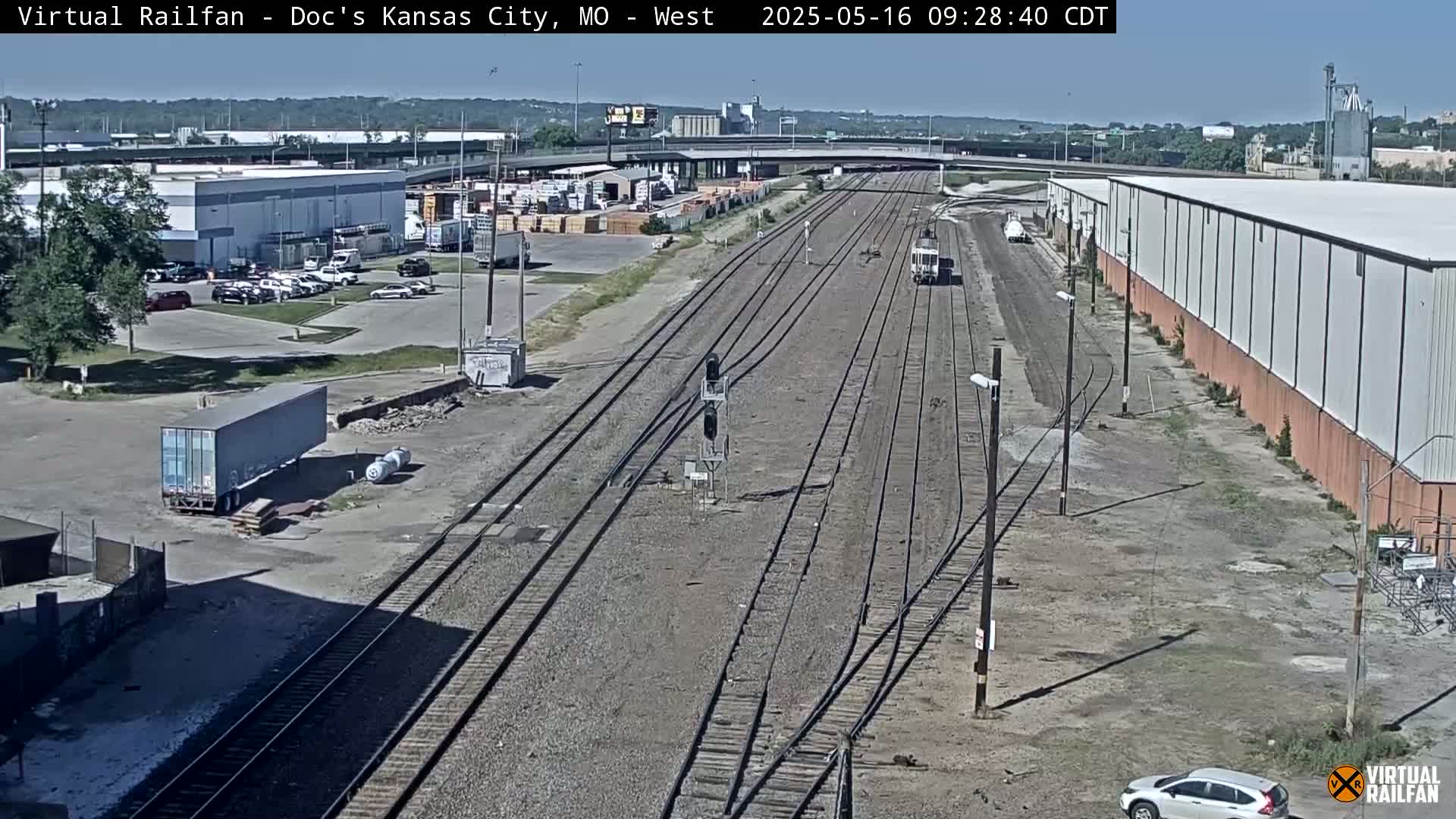 A high-angle, long shot on a sunny day shows several sets of train tracks extending into the distance, with buildings, cars, and a freight container visible on either side of the tracks.