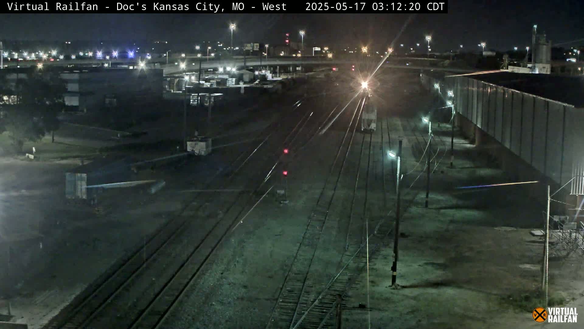 A nighttime view of a train yard with several railway tracks, industrial buildings, and a train approaching a signal.