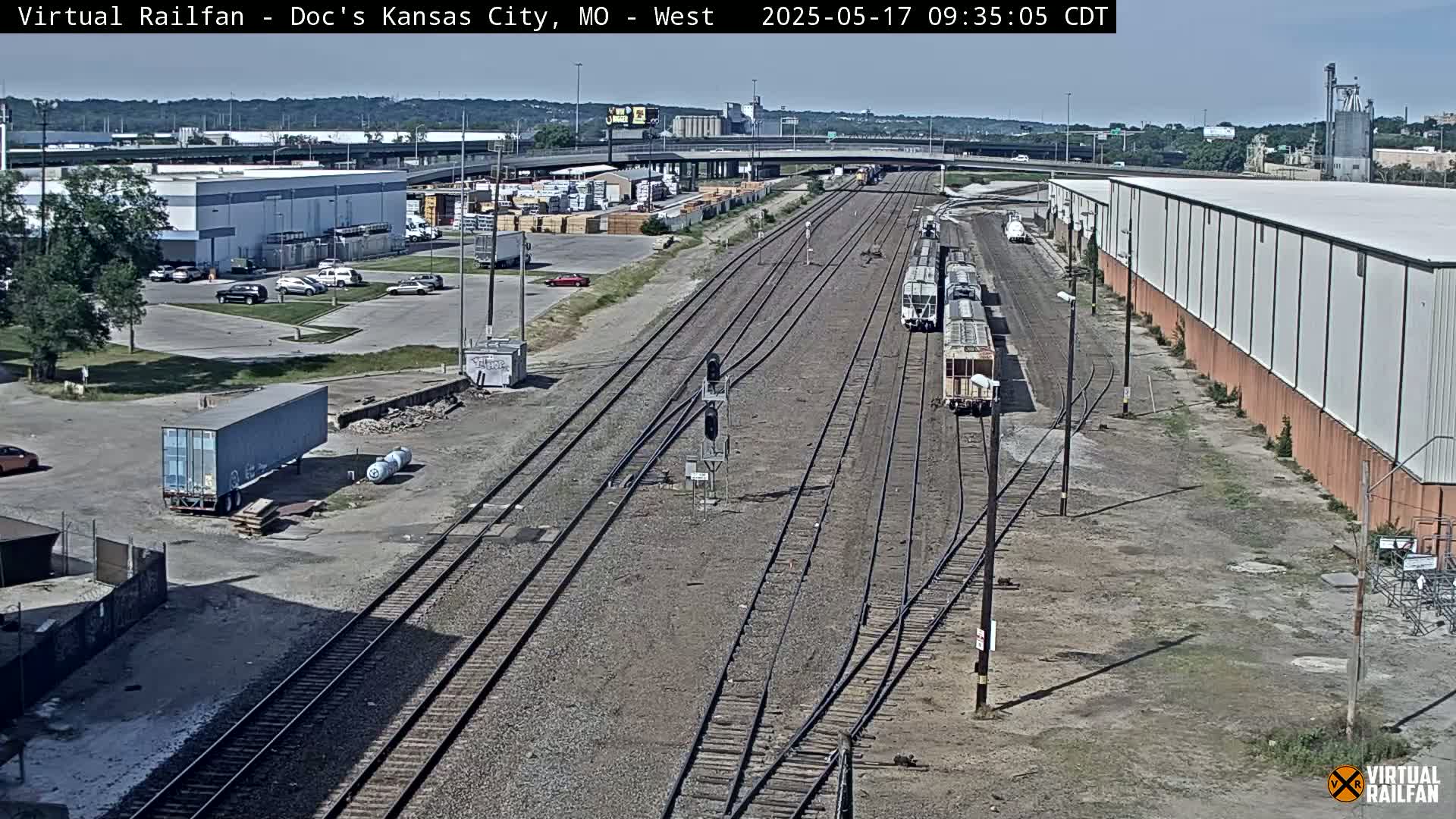 A high-angle, long shot on a sunny day shows several train cars stopped on railroad tracks next to industrial buildings and parking lots.
