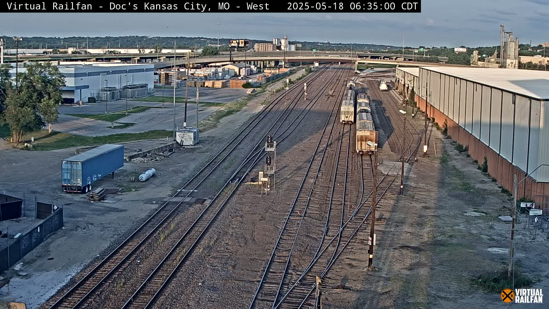 A high-angle, long shot of a rail yard with several train cars, industrial buildings, and a semi-trailer, under a partly sunny sky.
