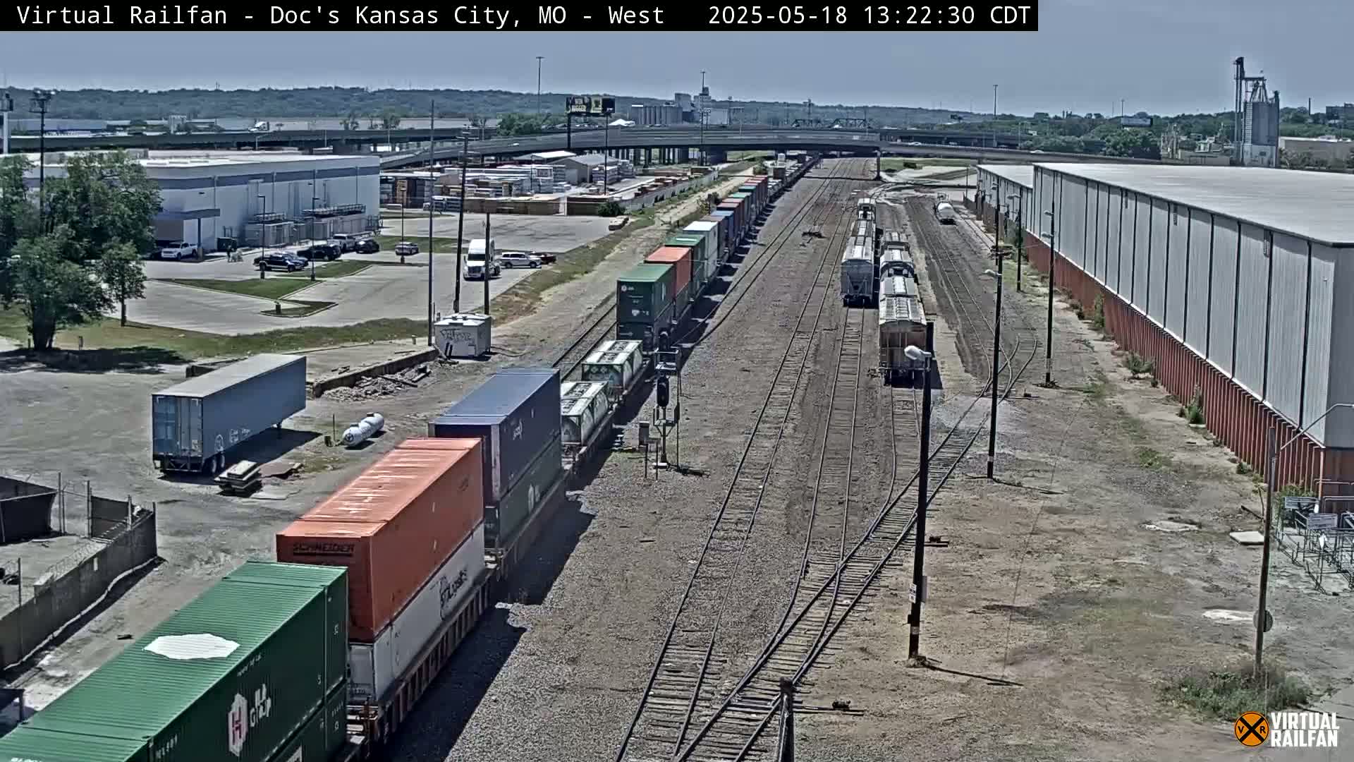 A long freight train carrying various cargo containers is visible on railroad tracks next to industrial buildings on a sunny day.
