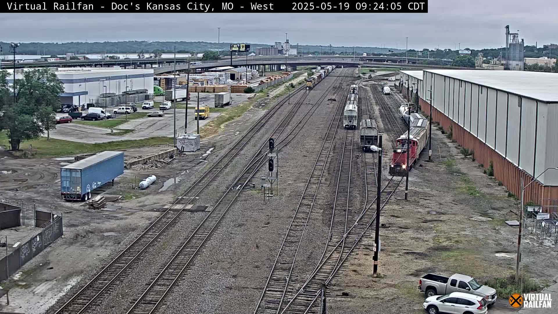 A railroad yard with several train cars and locomotives, surrounded by industrial buildings and parking lots under an overcast sky.