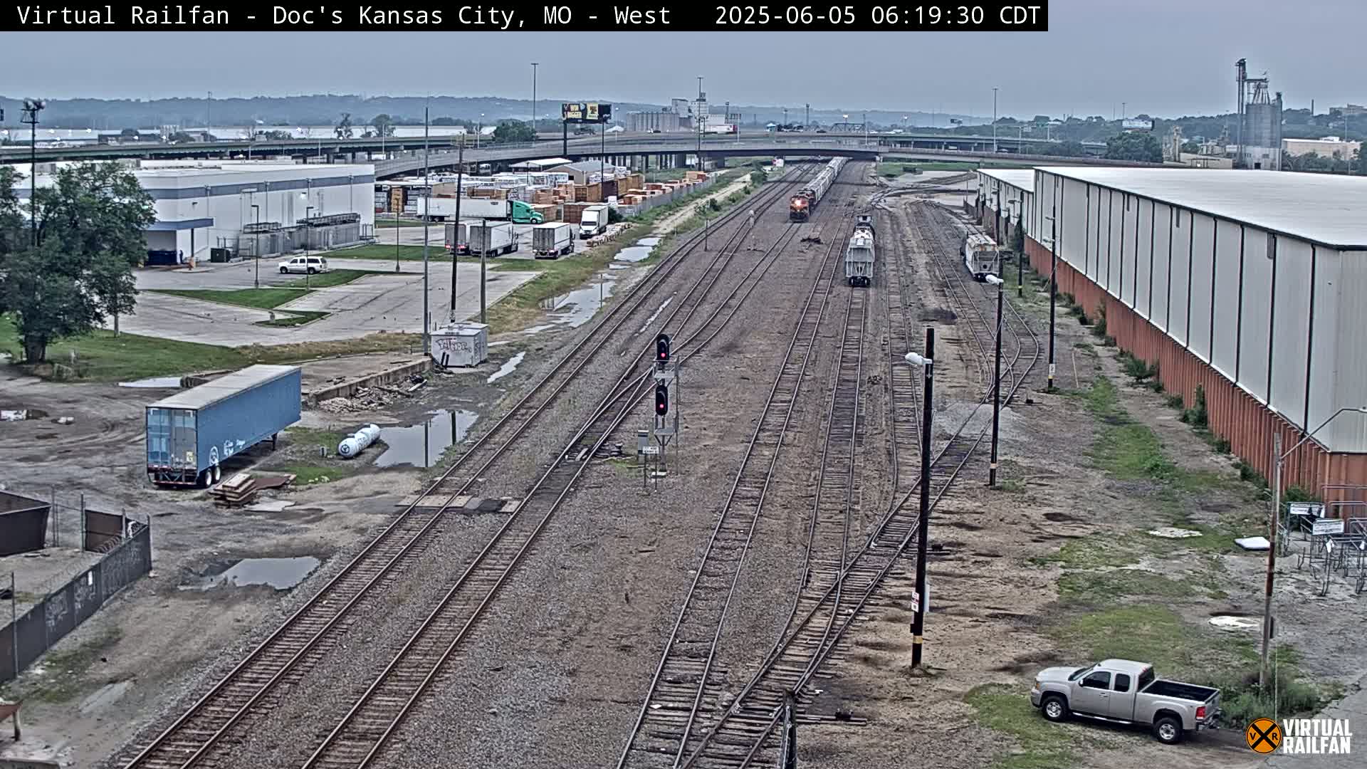 A high-angle, long shot of several train cars on railroad tracks, surrounded by industrial buildings and patchy vegetation under an overcast sky.