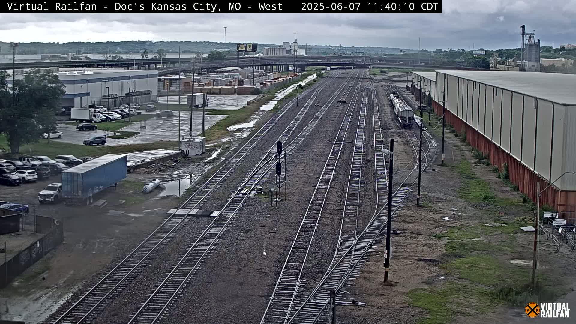 A high-angle view of railroad tracks and industrial buildings on an overcast day, with puddles of water visible on the ground.
