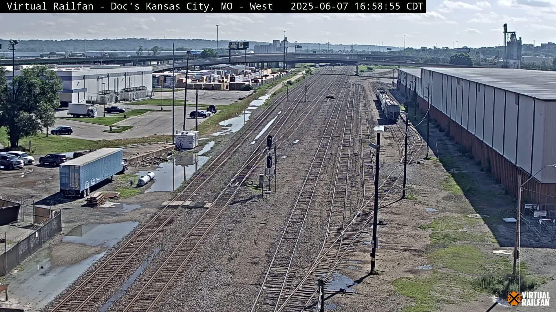 A mostly sunny view of a railway yard with several train tracks, industrial buildings, and puddles of water on the ground.