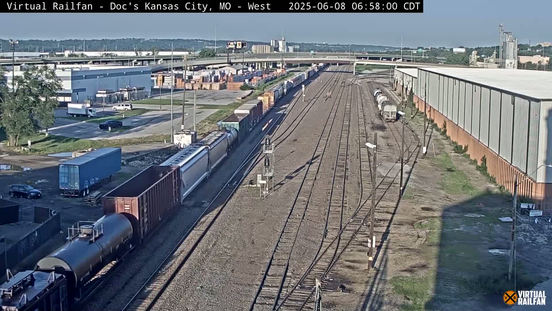 A long freight train is passing through a rail yard on a sunny day, near industrial buildings and roads.