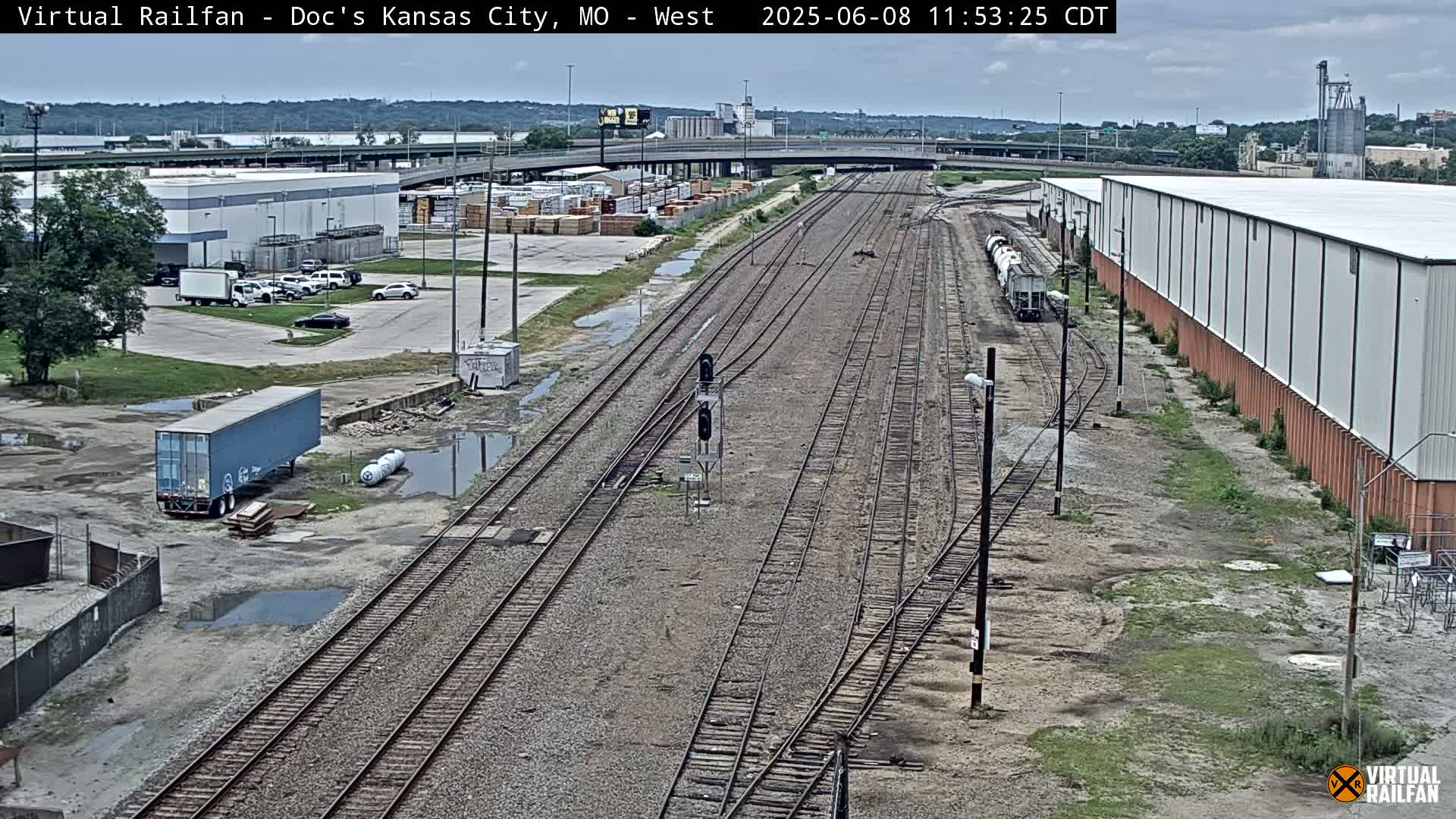 A high-angle view of a railway yard on an overcast day, showing several train tracks branching off from a main line, surrounded by industrial buildings and some puddles of water.