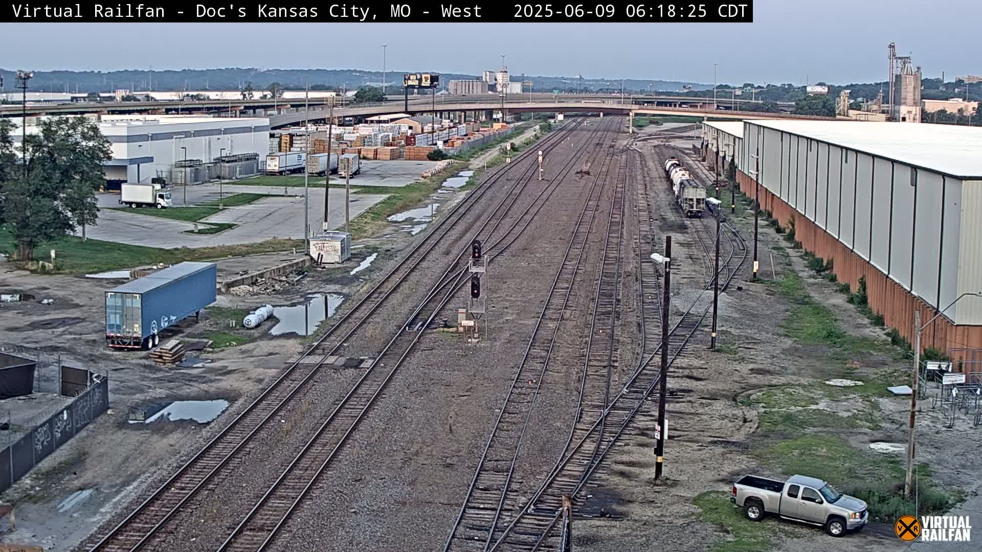 A high-angle, long shot of railroad tracks and industrial buildings on an overcast day, with several trucks and a train visible.