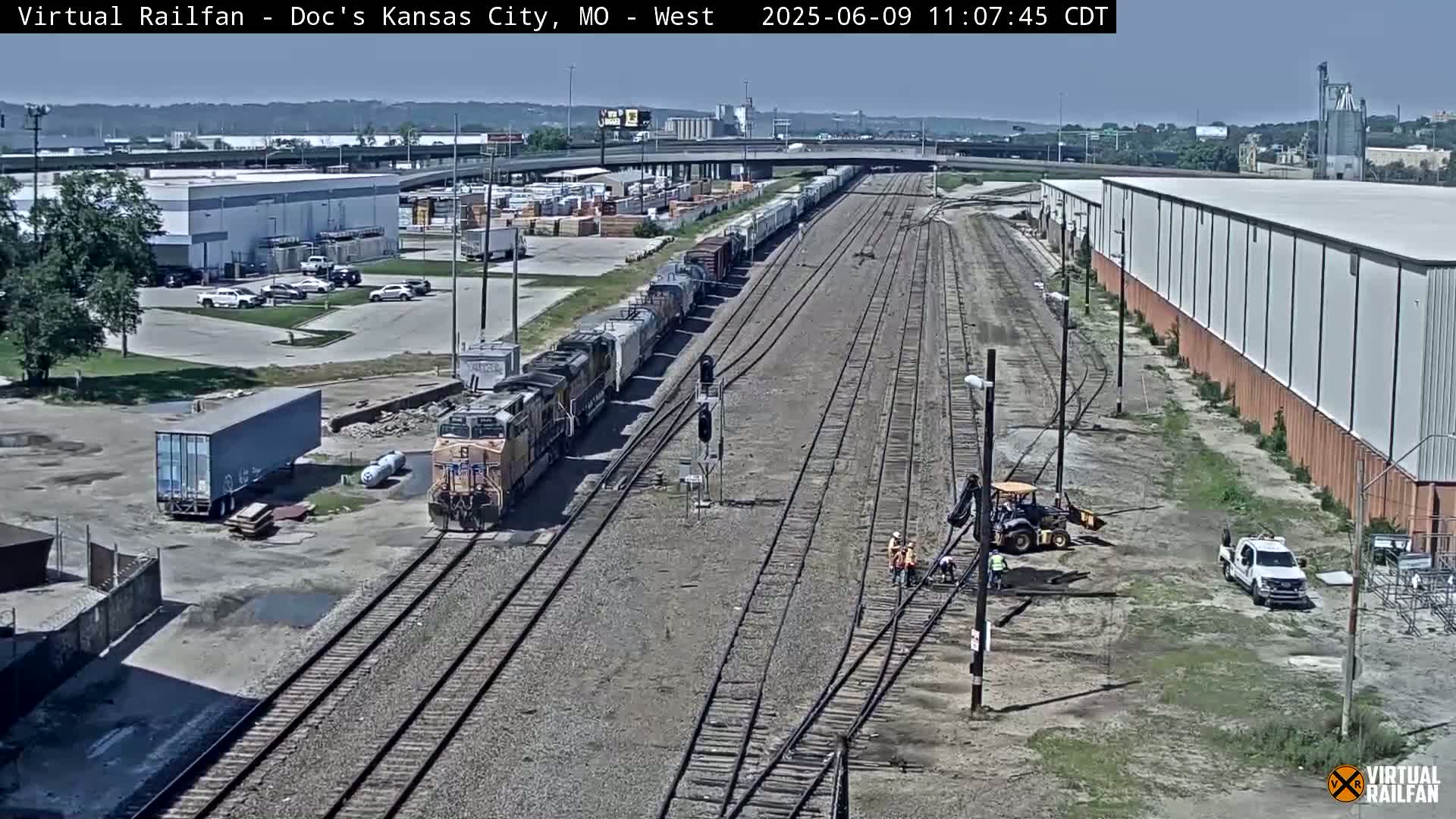 A long freight train is passing through a rail yard on a sunny day, with several workers and pieces of industrial equipment visible nearby.