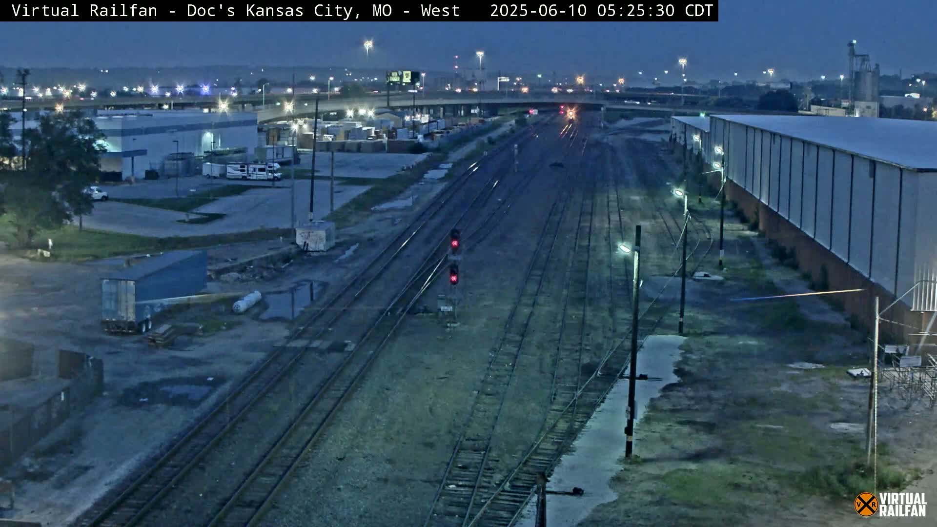 A nighttime view of a railroad yard with several train tracks, industrial buildings, and a few vehicles, under a dark sky.
