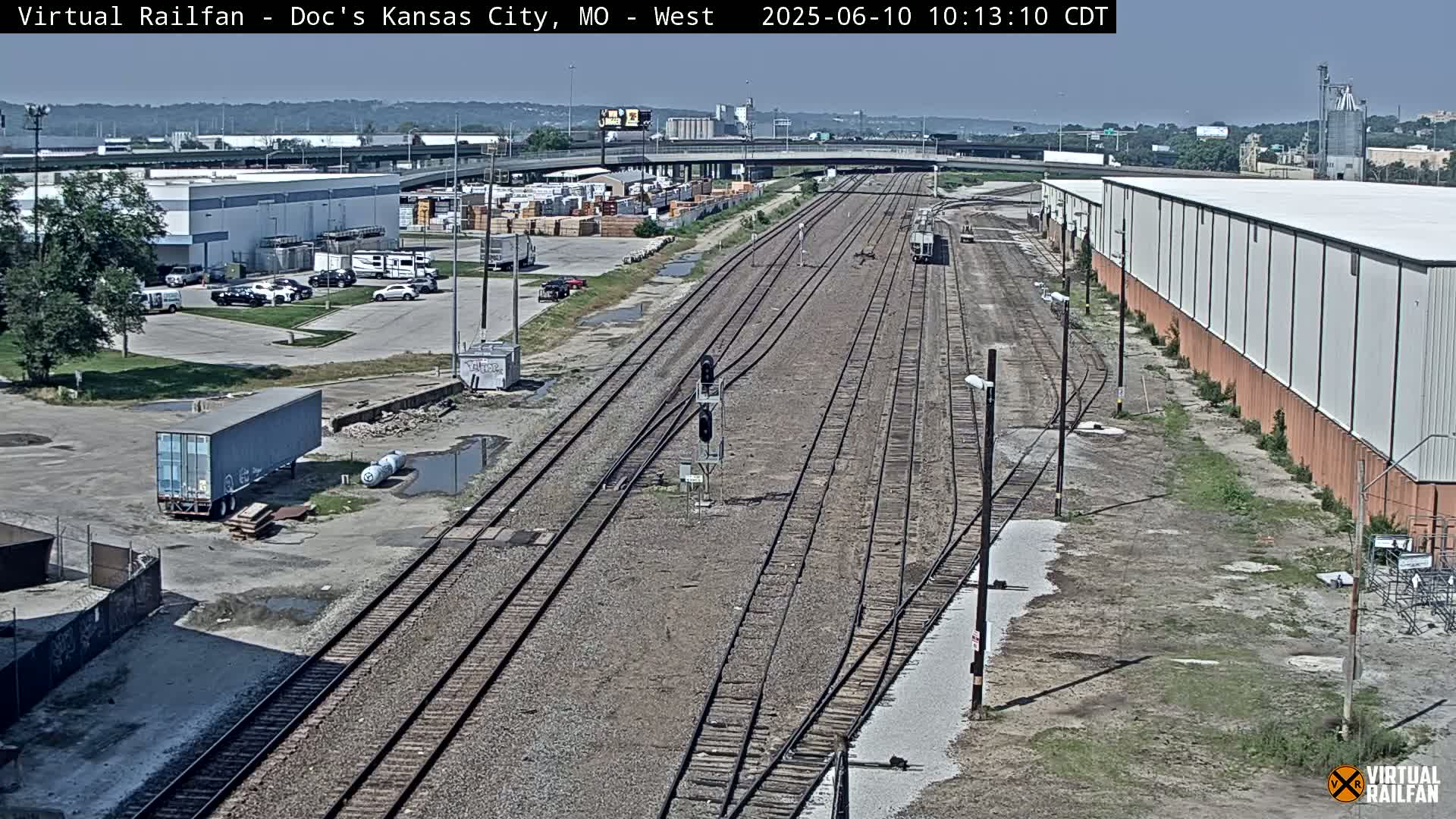 A high-angle view of a railway track system on a sunny day, surrounded by industrial buildings and parking lots.
