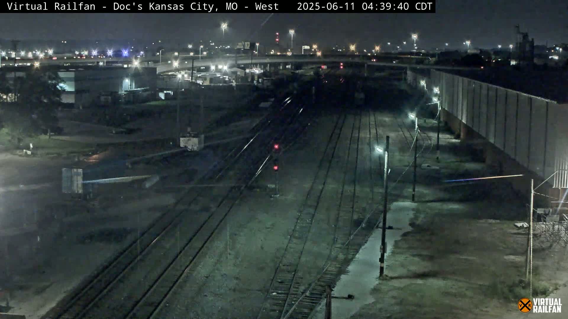 A nighttime view of railroad tracks extending into the distance, alongside industrial buildings and a highway under a clear sky.