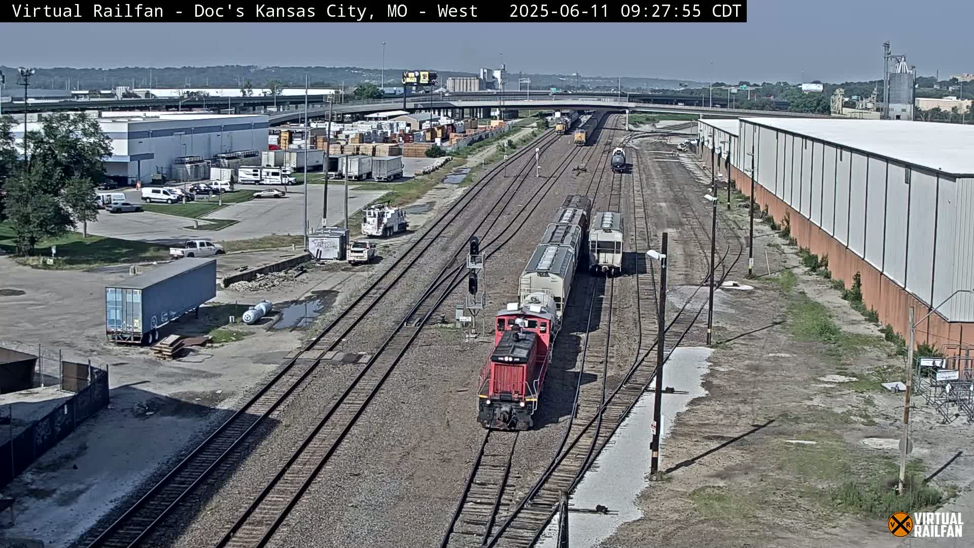 A red locomotive pulls a train of freight cars along railroad tracks in a sunny industrial area.