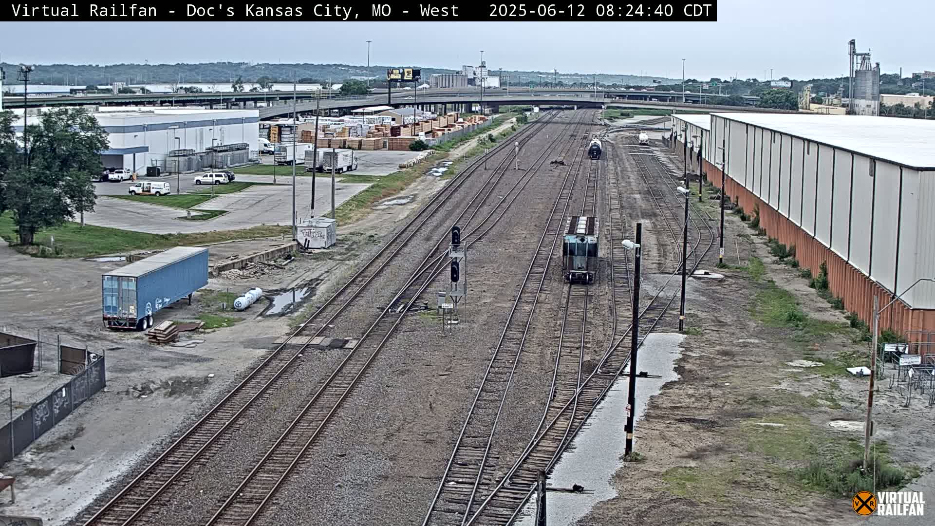 A high-angle view of a railway yard with several train cars and surrounding industrial buildings under overcast skies.