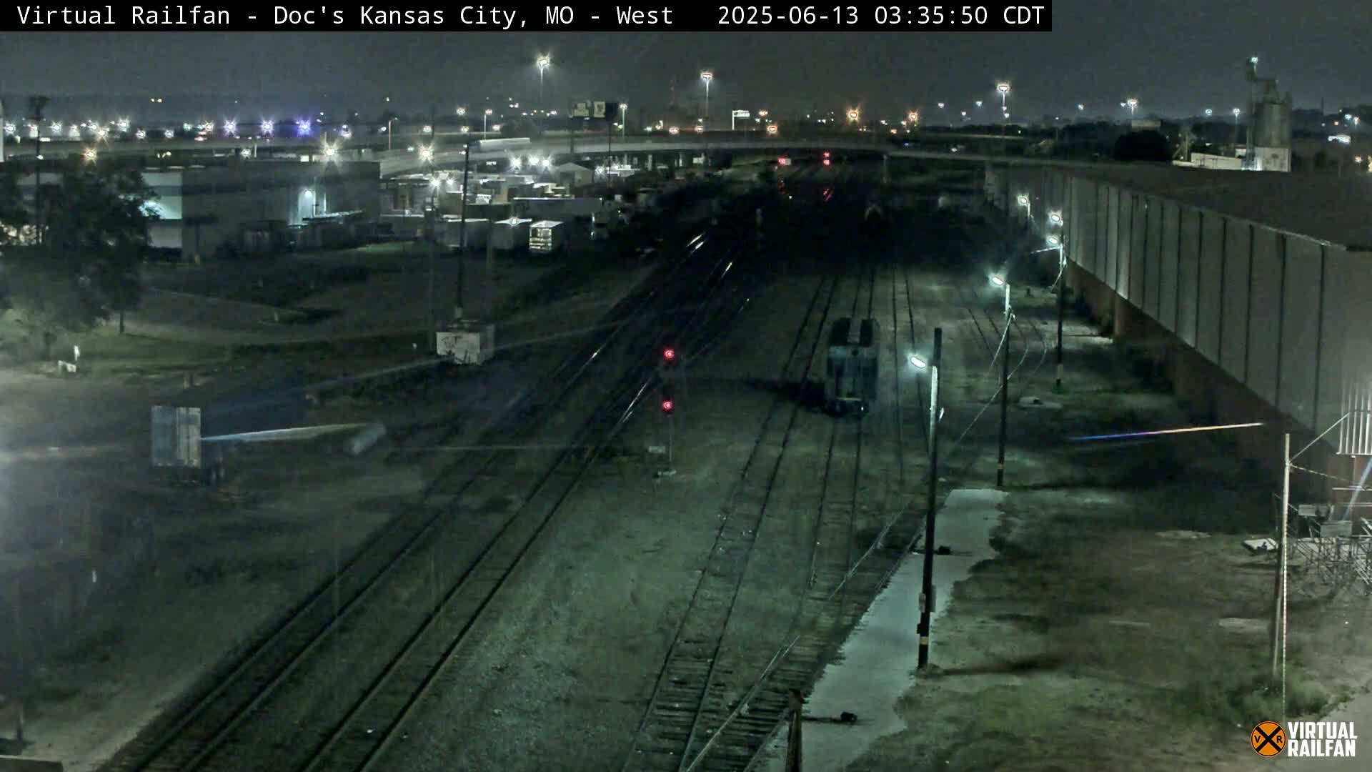 A nighttime view of railroad tracks and industrial buildings under clear skies, with a single locomotive visible on the tracks.