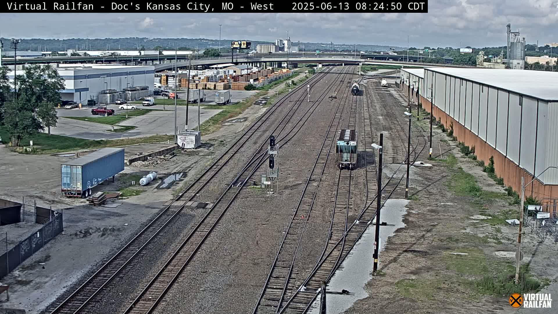 A mostly cloudy day shows a train on a rail yard with several industrial buildings and parking lots visible,  surrounded by patchy grass.