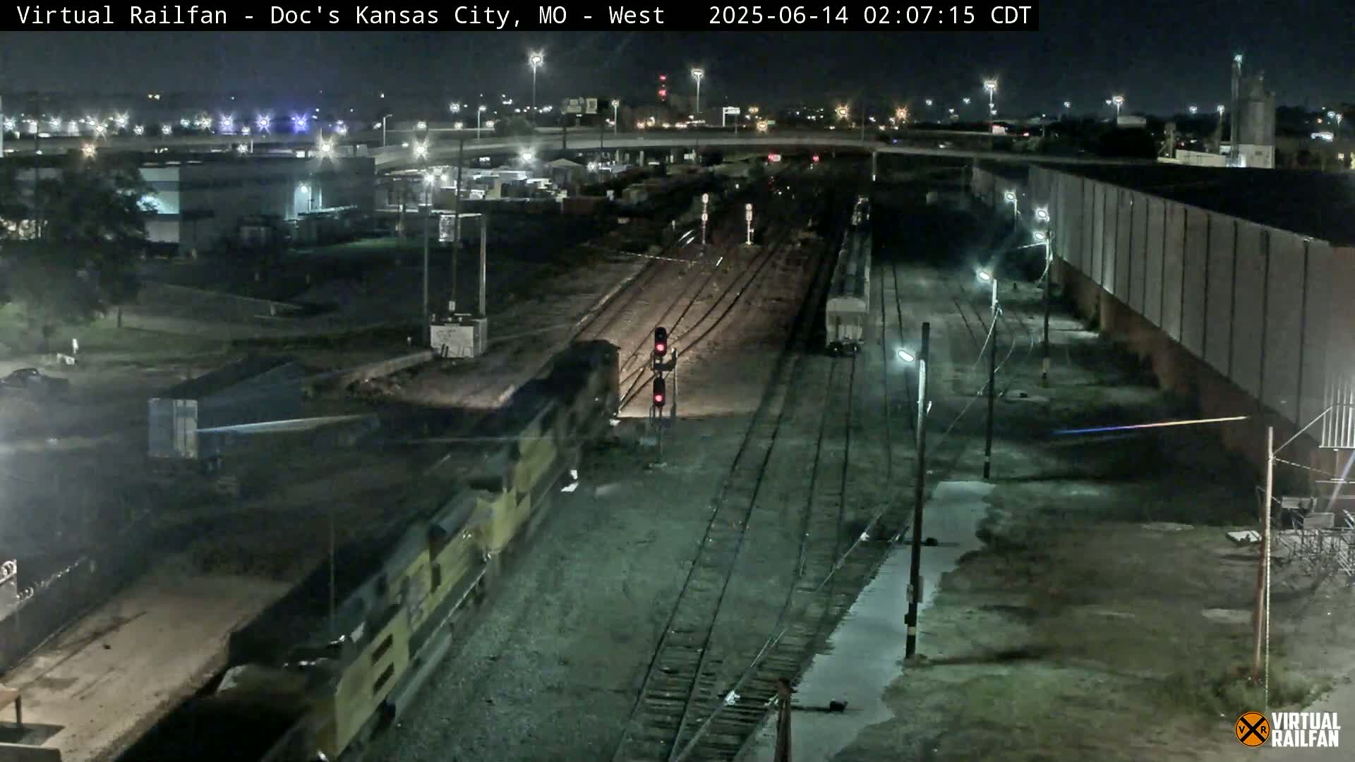 A nighttime view of a freight train approaching a signal at a rail yard.
