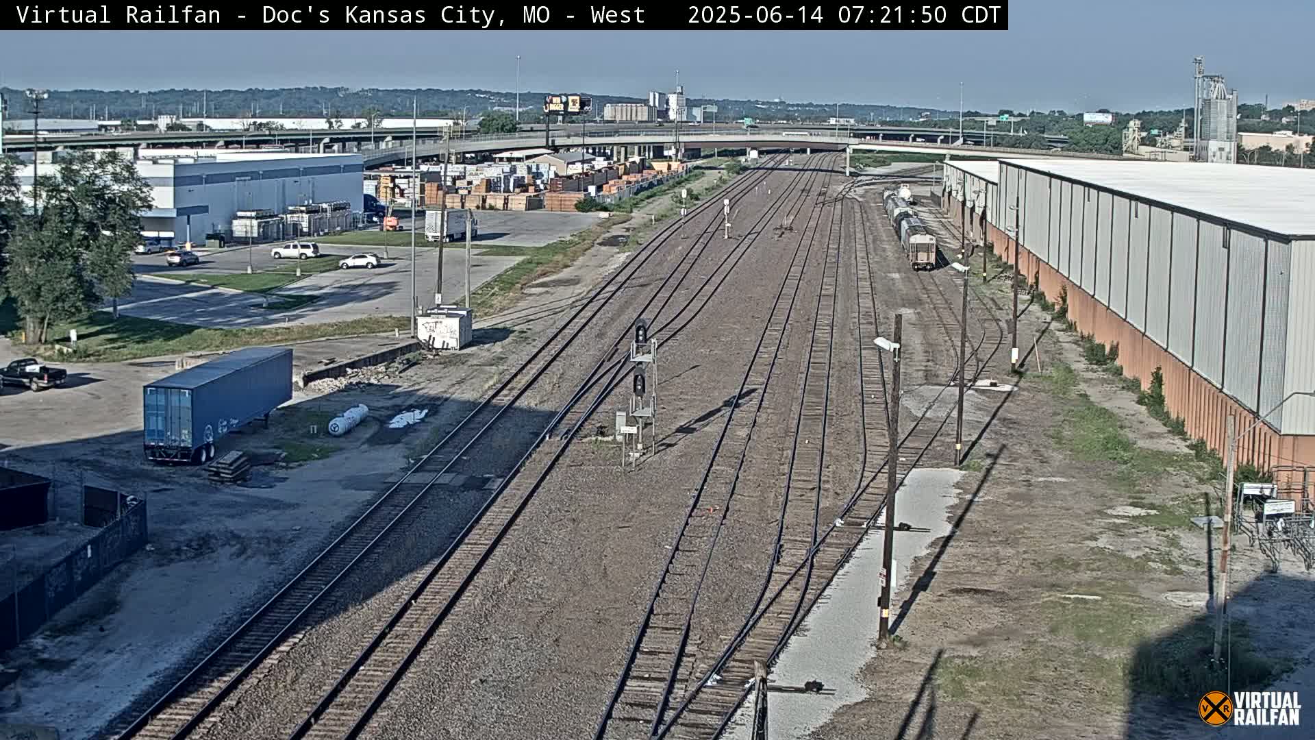 A high-angle, long shot of a railway yard on a sunny day, with several train cars visible, industrial buildings, and vehicles in a parking lot.